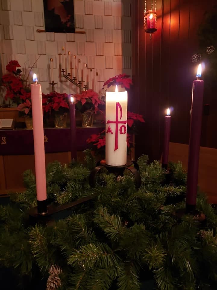 The Advent wreath at Immanuel, three purple candles and one rose lit around the white Christ candle marked with a Chi-Rho, set among poinsettias