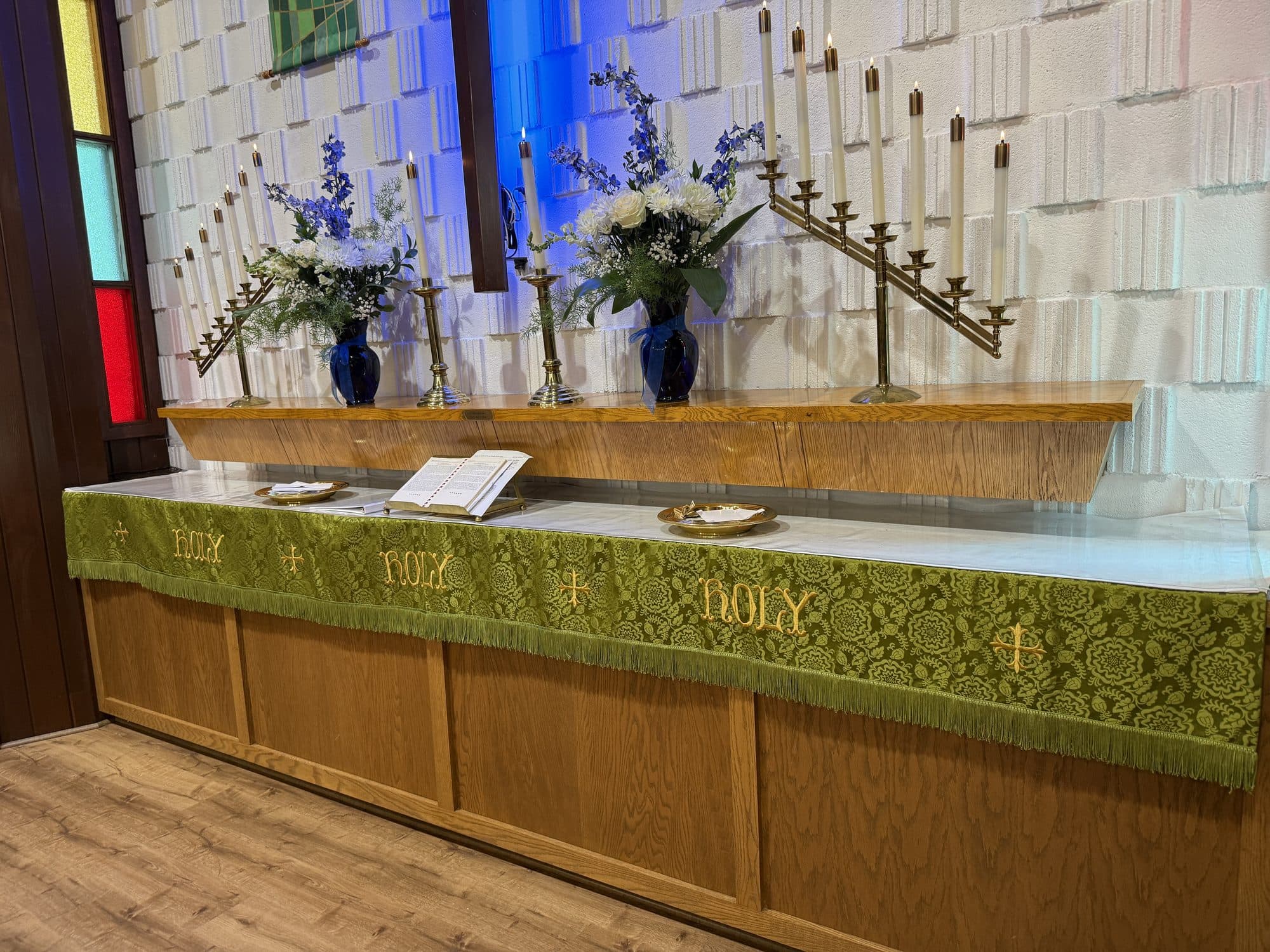 The altar dressed in green paraments embroidered HOLY, HOLY, HOLY in gold, with brass candelabra and blue and white flowers behind it
