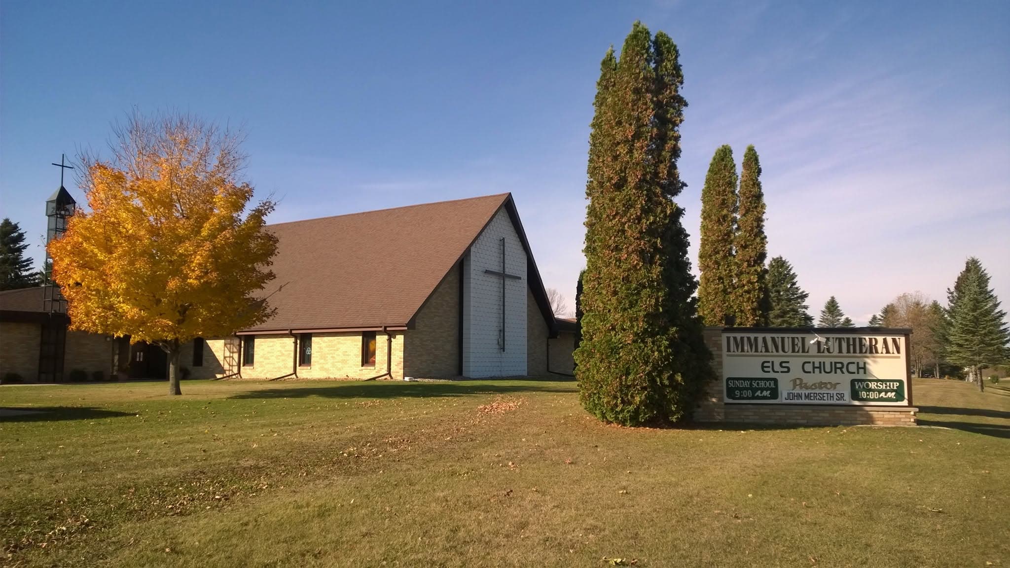 Immanuel Lutheran Church in autumn — brick walls, a cross on the gable, a bell tower, and a maple in full color beside the roadside sign