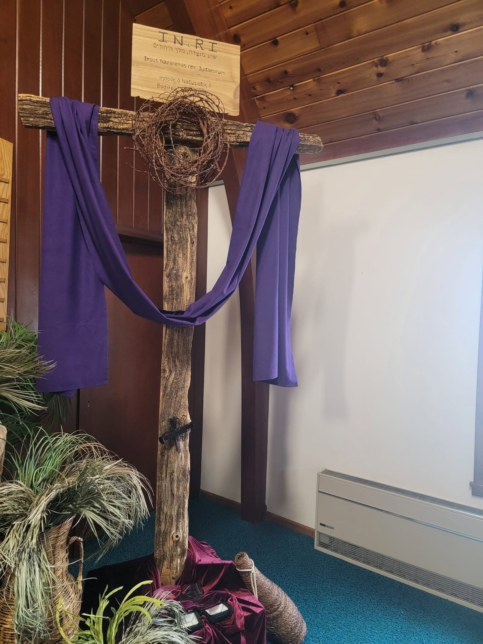 A rough timber cross in the sanctuary draped in purple, with a crown of thorns and an INRI placard lettered in Hebrew, Latin, and Greek