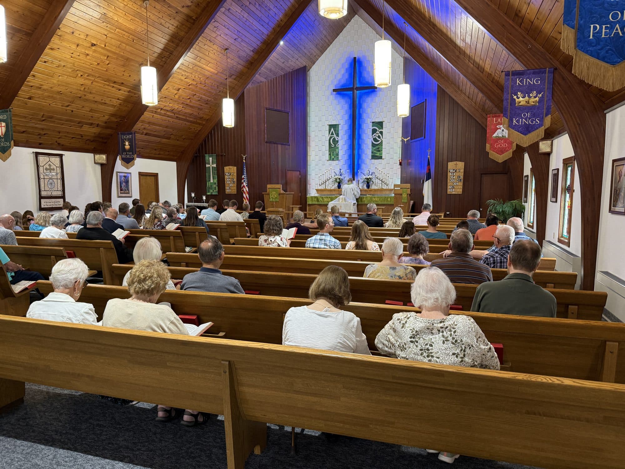 The congregation in the pews on a Sunday morning, facing the altar and the lit cross beneath a timber-vaulted ceiling hung with banners